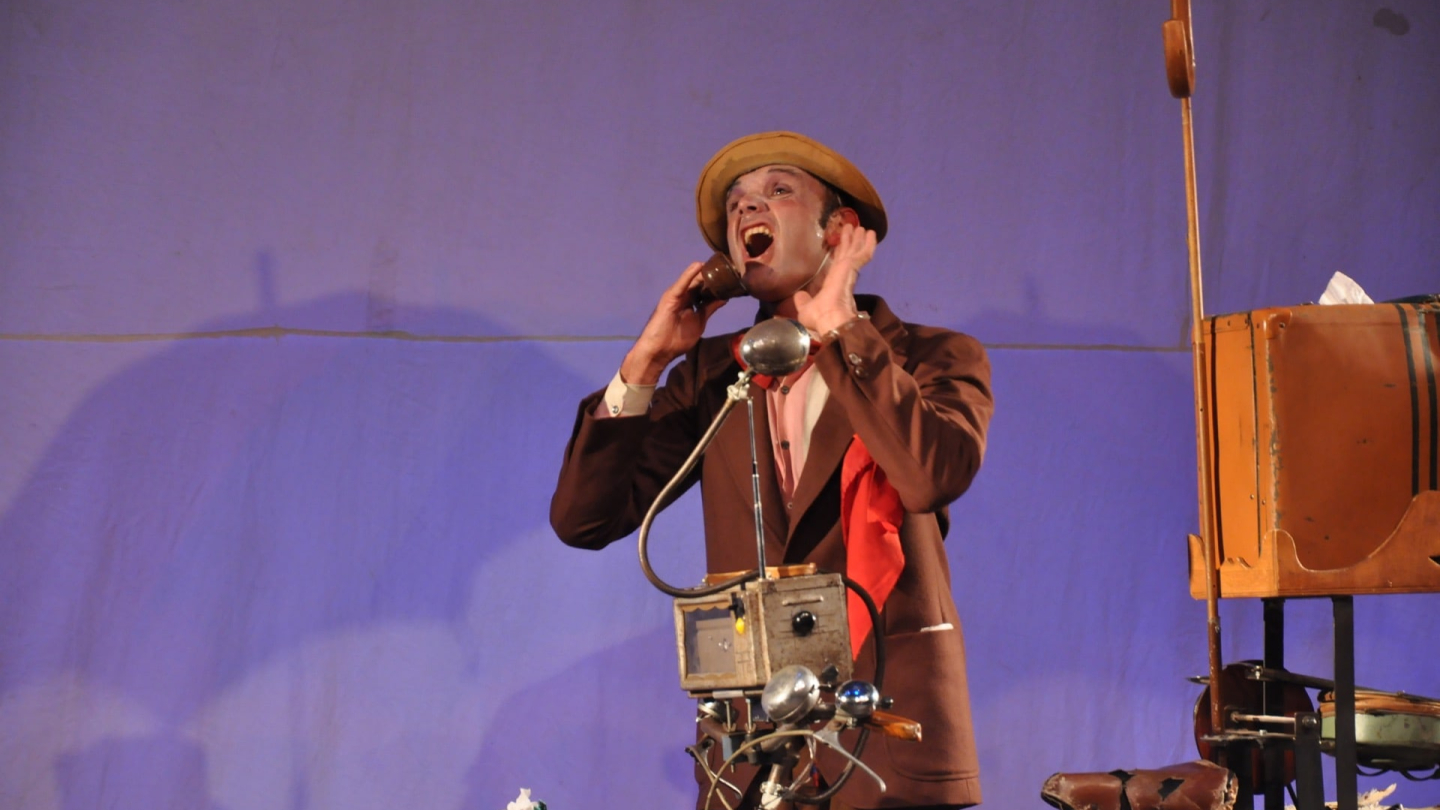 Fotografia de um ator caracterizado com terno marrom e chapéu, expressando surpresa ou grito enquanto utiliza um dispositivo cênico acoplado a uma bicicleta em frente a um fundo azul