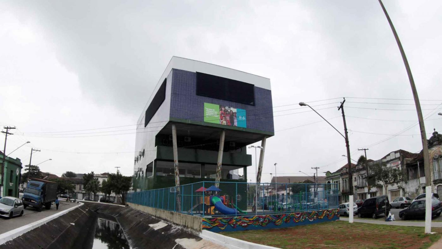 Uma construção moderna e geométrica com parte de sua estrutura elevada por pilares se destaca em uma área urbana, cercada por um playground infantil, um canal e casas antigas sob um céu nublado