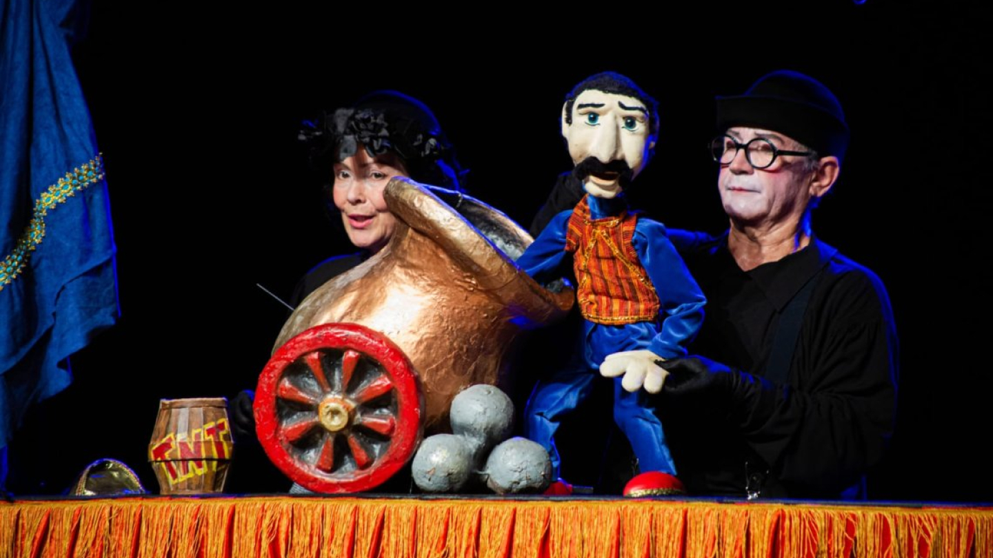 Dois artistas manipulam um boneco de bigode e um pequeno canhão dourado em uma apresentação de teatro de bonecos com temática circense