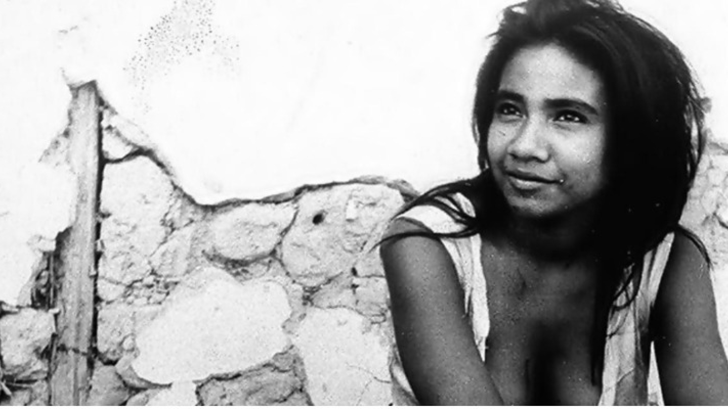 Retrato em preto e branco de uma mulher jovem com olhar sereno, posicionada à frente de uma parede rústica de pedras e barro