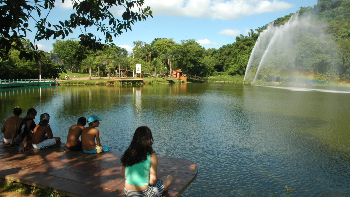 Um grupo de jovens descansa em um deck de madeira à beira da Lagoa da Saudade, observando as fontes de água que formam um arco-íris sob o céu ensolarado