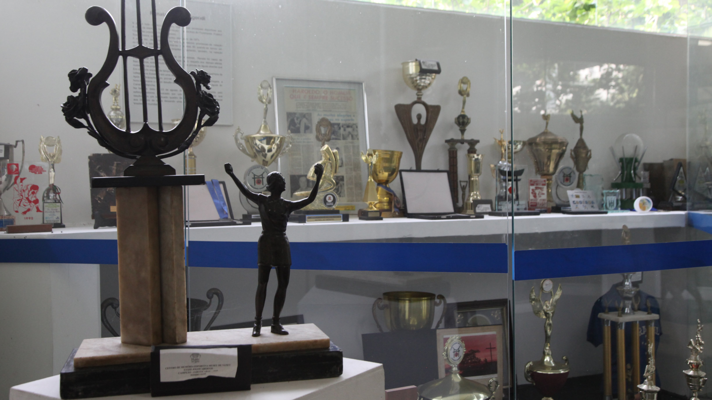 Uma vitrine de vidro exibe diversos troféus e medalhas esportivas, com destaque para uma estatueta de uma atleta em primeiro plano