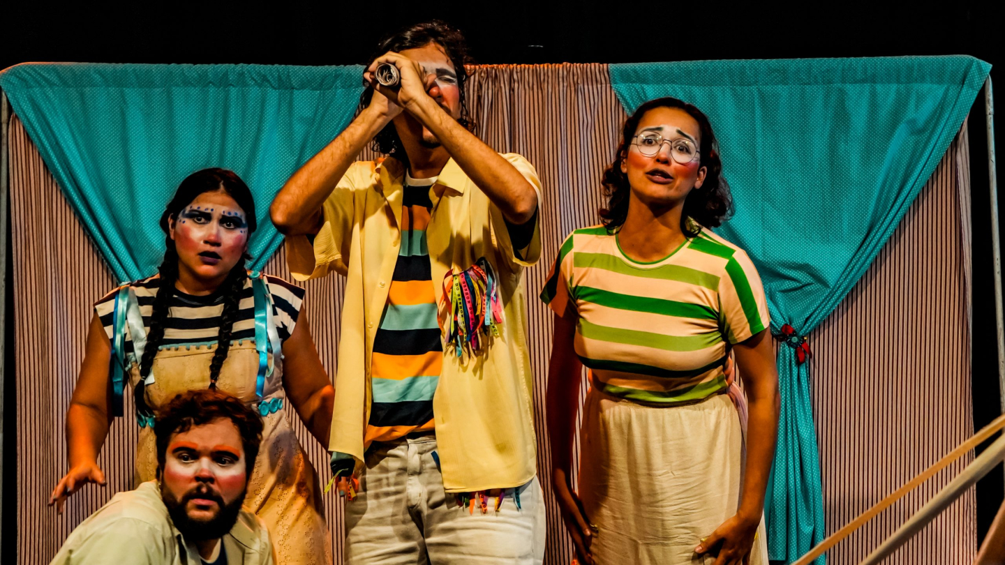Quatro atores com maquiagem artística e figurinos coloridos encenam a peça teatral "Onde Está o Guará?" em frente a uma cortina listrada, com um dos personagens utilizando um monóculo improvisado para olhar à distância
