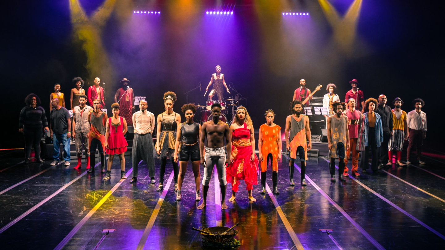 Fotografia do espetáculo "ProtAGÔnistas - O Movimento Negro no Picadeiro", mostrando um numeroso elenco de artistas negros posicionado no palco sob luzes cênicas