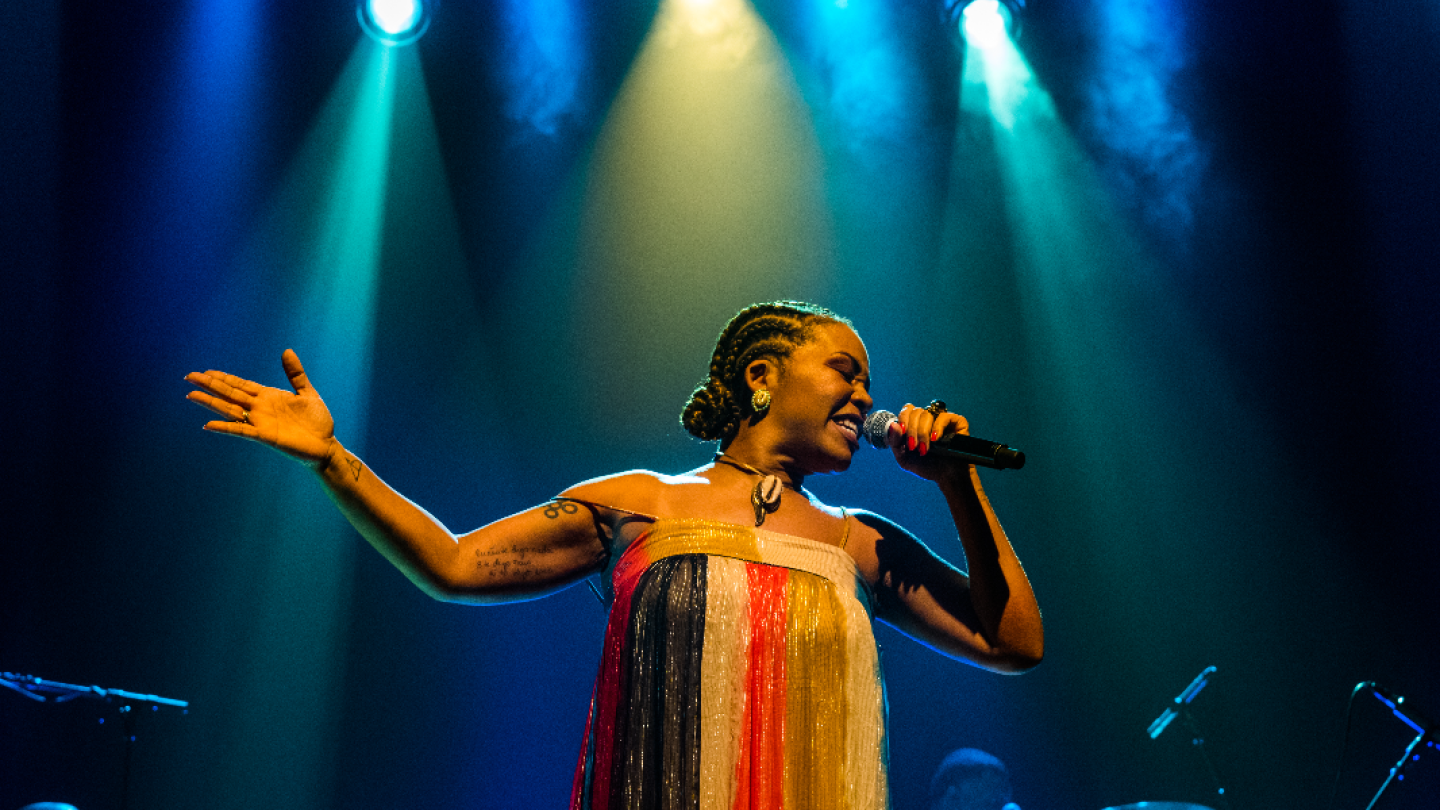 Uma cantora com tranças nagô e vestido colorido se apresenta intensamente sob luzes de palco azuladas e amareladas