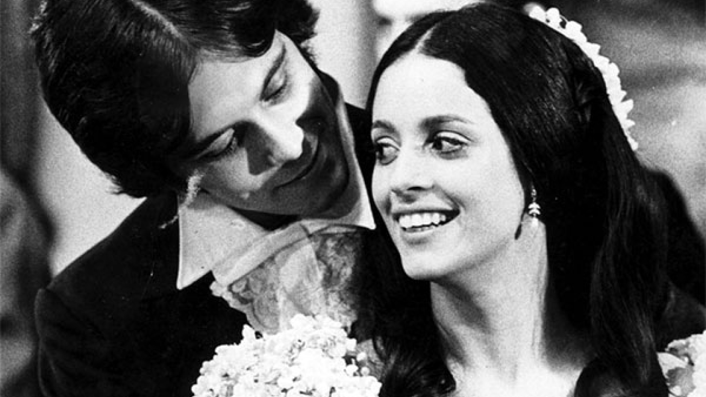 Uma fotografia em preto e branco captura um casal jovem com trajes de época, onde o homem olha carinhosamente para a mulher enquanto ela sorri