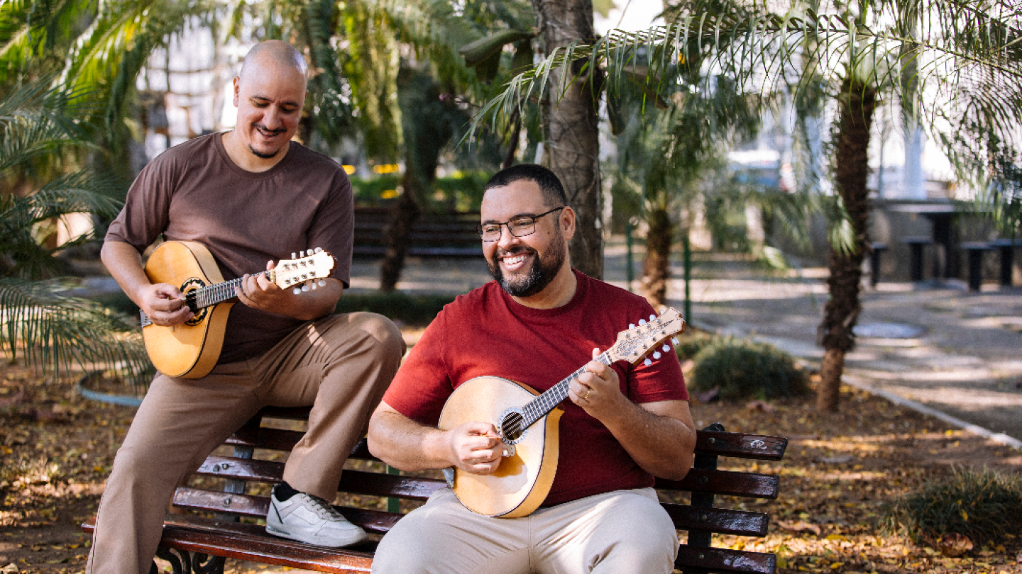 Dois músicos aparecem sorridentes sentados em um banco de praça, cercados por palmeiras, enquanto tocam bandolins de dez cordas