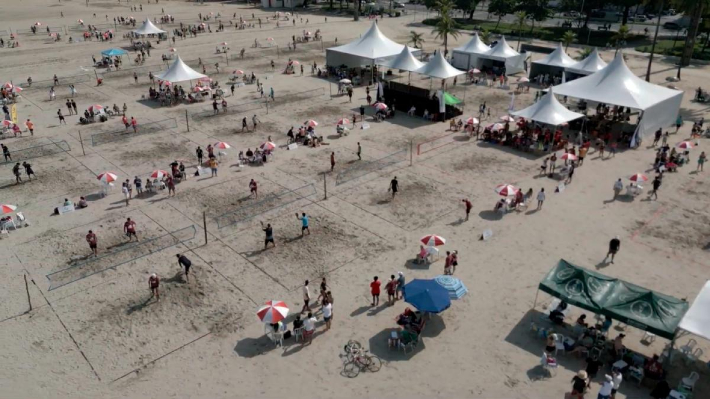 Vista aérea de um torneio de Beach Tennis em uma praia movimentada, apresentando diversas quadras montadas na areia e estruturas de tendas brancas para o público e organização