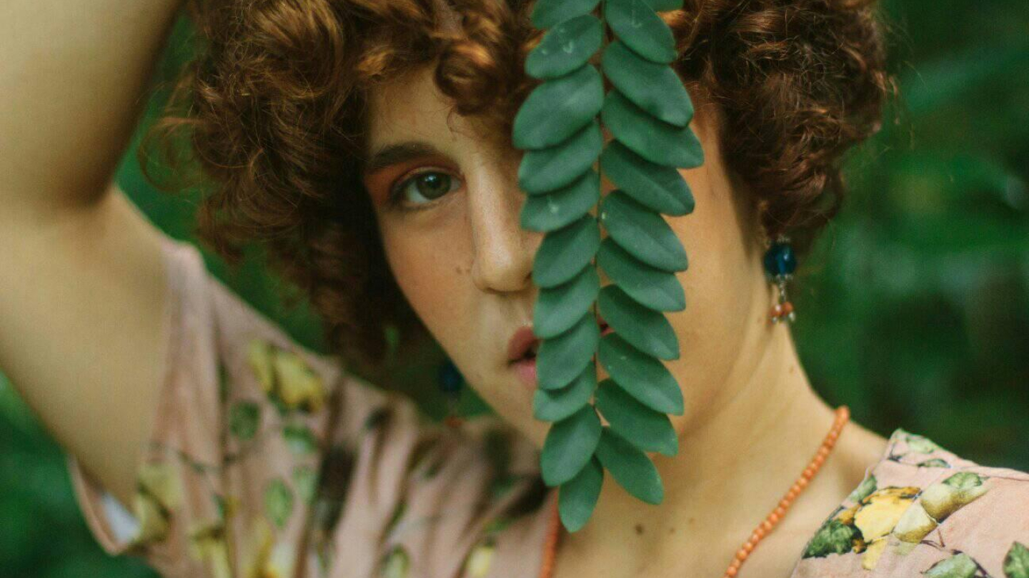 Uma pessoa jovem com cabelos cacheados posa entre folhagens, segurando um ramo de folhas que encobre parcialmente o seu rosto