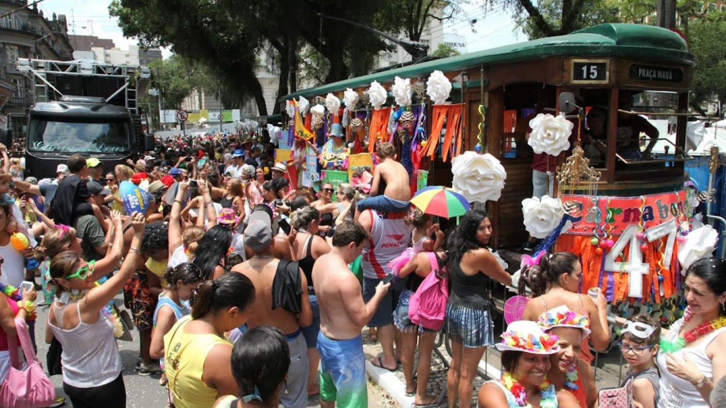 Uma multidão festiva e colorida celebra o Carnaval ao redor de um bonde histórico decorado que exibe o letreiro "Carnabonde"