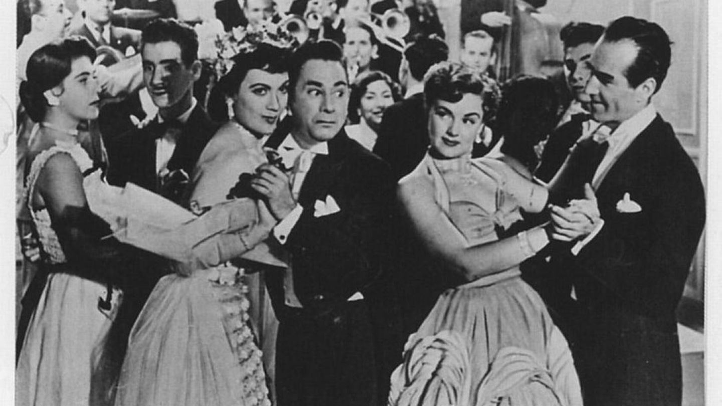 Uma cena em preto e branco do filme brasileiro Carnaval Atlântida (1952) mostra casais elegantemente vestidos dançando em um salão festivo, com destaque para os atores Oscarito e Eliana Macedo ao centro