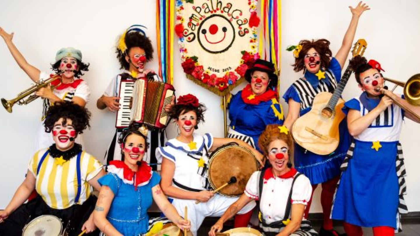 Um grupo de palhaças sorridentes e coloridas posa com instrumentos musicais sob um estandarte do grupo "Sampalhaças"