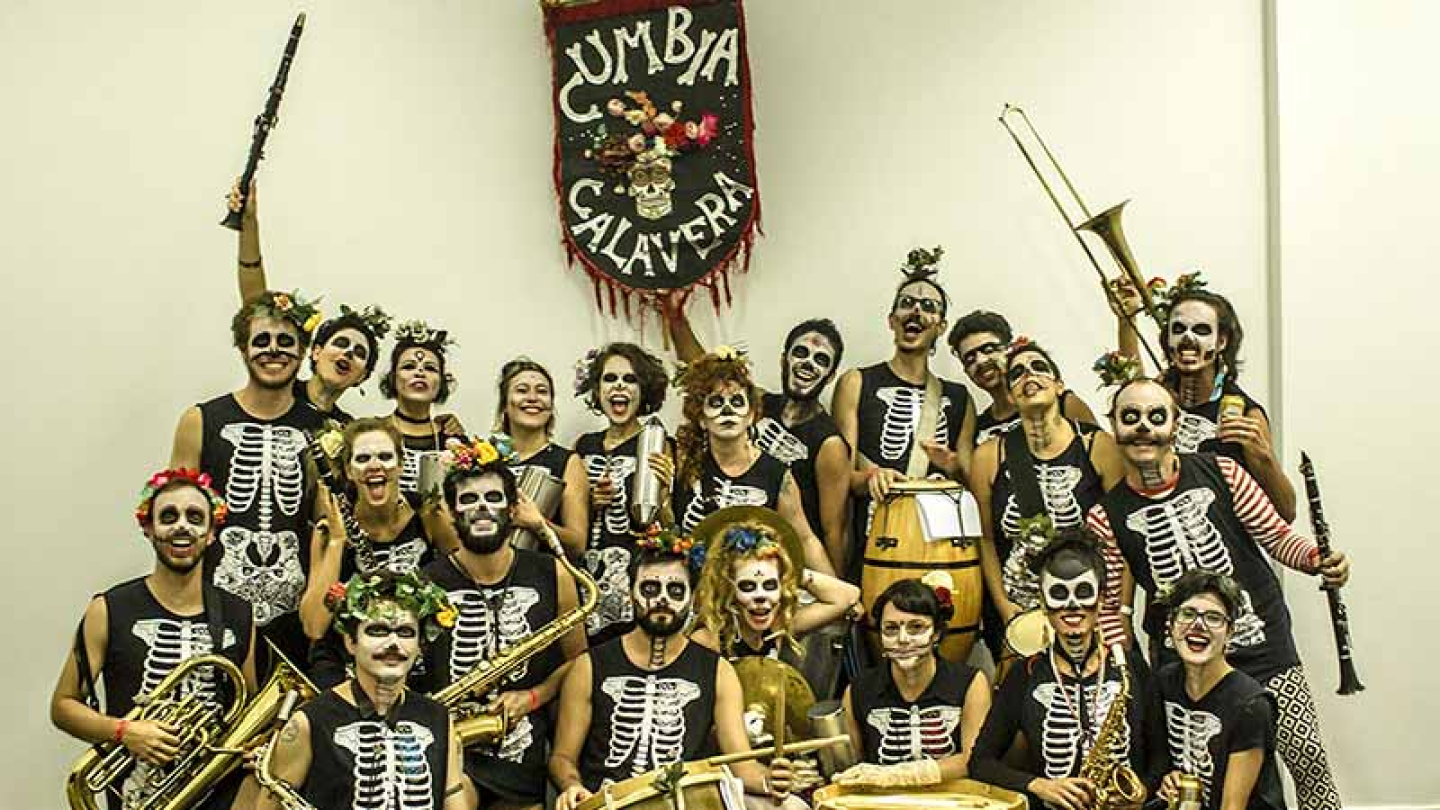 Os integrantes da banda Cumbia Calavera posam sorridentes com seus instrumentos, vestindo roupas de esqueleto e maquiagens de caveira mexicana sob o estandarte do grupo