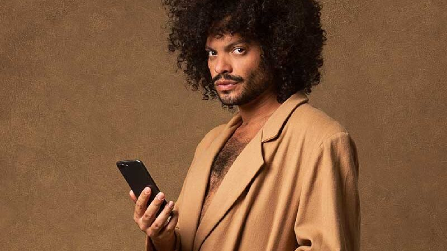 Retrato de estúdio do cantor Diego Moraes com cabelo estilo black power, vestindo um sobretudo bege e segurando um smartphone contra um fundo de textura marrom