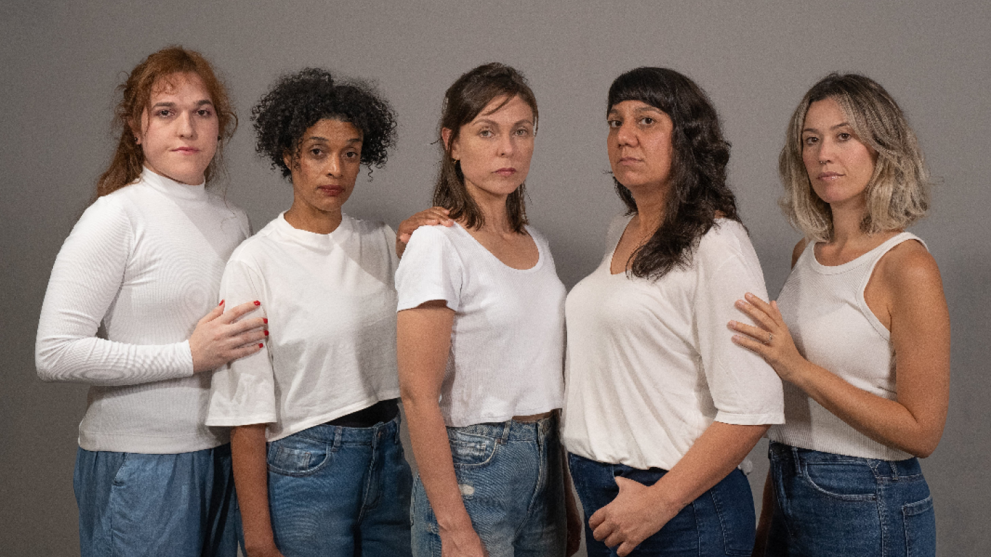 Cinco mulheres com expressões sérias posam lado a lado contra um fundo cinza, vestindo blusas brancas e calças jeans, com algumas apoiando as mãos nos ombros umas das outras