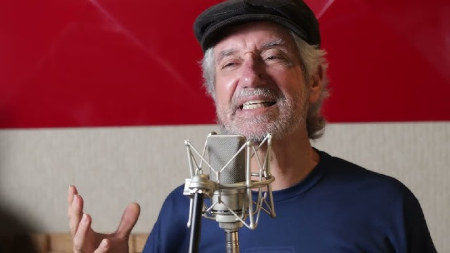 O músico Edu Quintana aparece sorrindo e gesticulando enquanto canta em frente a um microfone de estúdio profissional