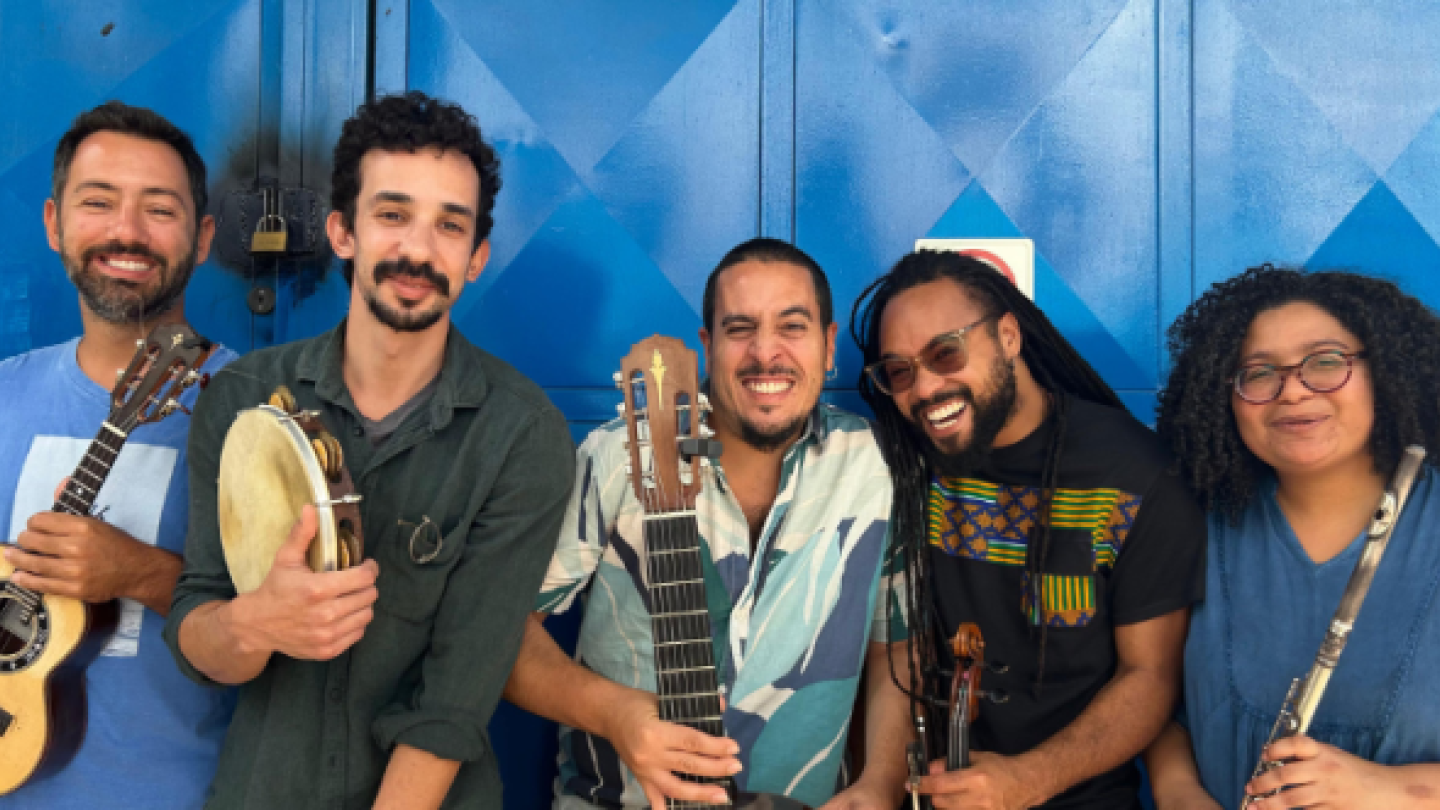 Fotografia de cinco músicos sorridentes do grupo Fios de Choro posando em frente a um portão azul, segurando instrumentos como cavaquinho, pandeiro, violão de sete cordas e flauta transversal