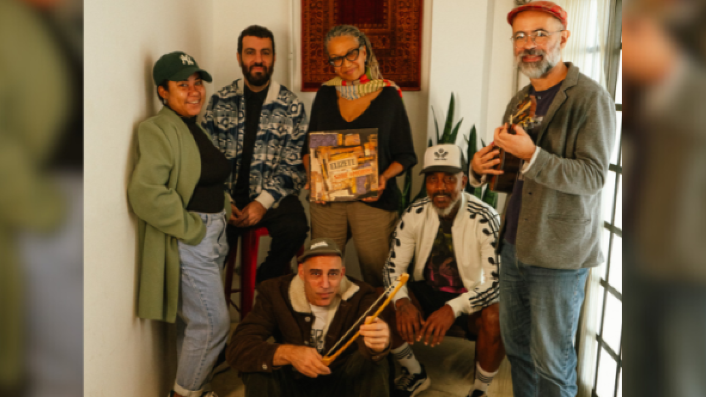A fotografia mostra um grupo de seis músicos, incluindo a cantora Juçara Marçal, posando em um ambiente interno com instrumentos musicais e um disco de vinil