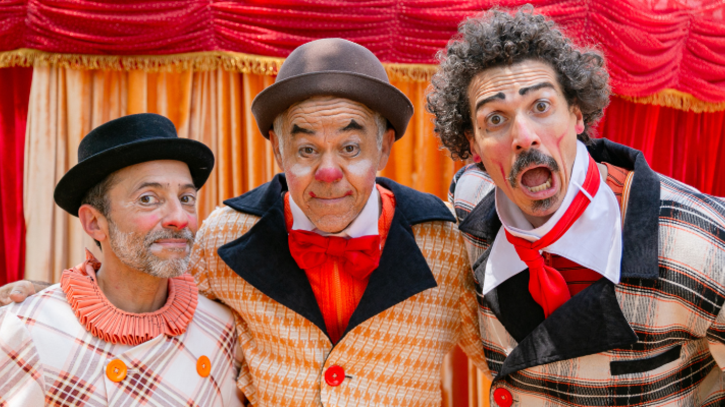 Três palhaços com expressões cômicas e roupas coloridas posam abraçados em frente a uma cortina de circo vermelha