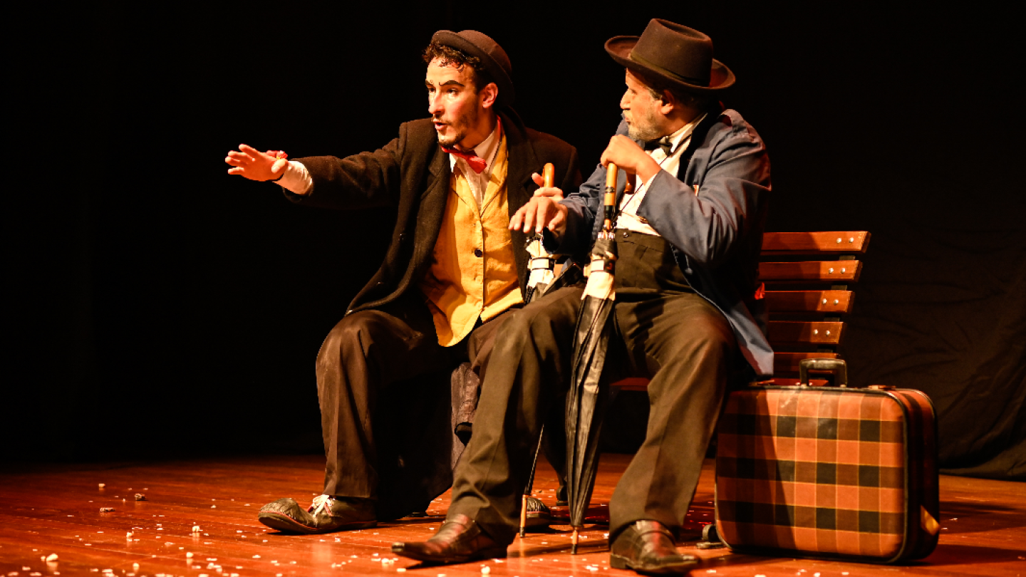 Dois atores caracterizados com chapéus e paletós contracenam sentados em um banco de praça sobre um palco de teatro