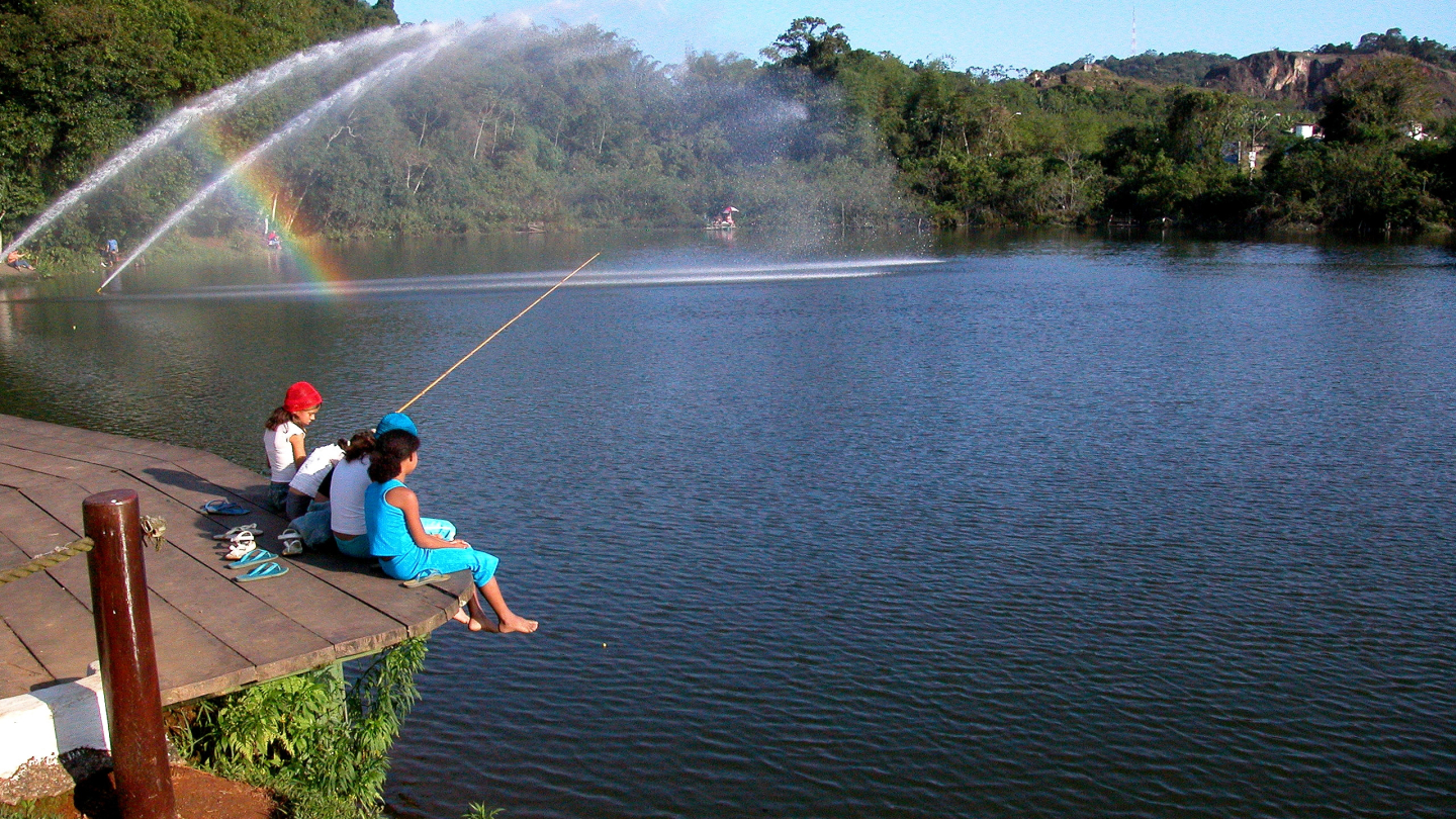 Crianças e jovens pescam e relaxam em um deck de madeira à beira da Lagoa da Saudade, onde fontes de água criam um arco-íris