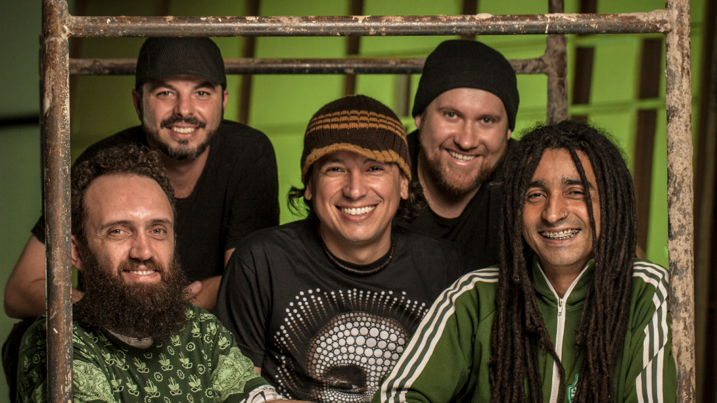Cinco homens sorridentes, incluindo um com dreadlocks e outro com barba longa, posam juntos atrás de uma estrutura metálica simples