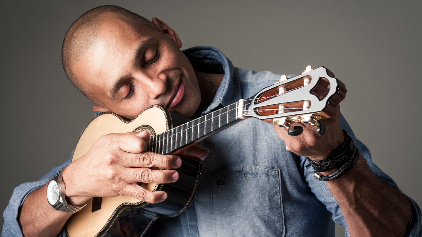 Um homem de cabeça raspada e olhos fechados encosta o rosto carinhosamente em um cavaquinho, demonstrando uma conexão profunda com o instrumento