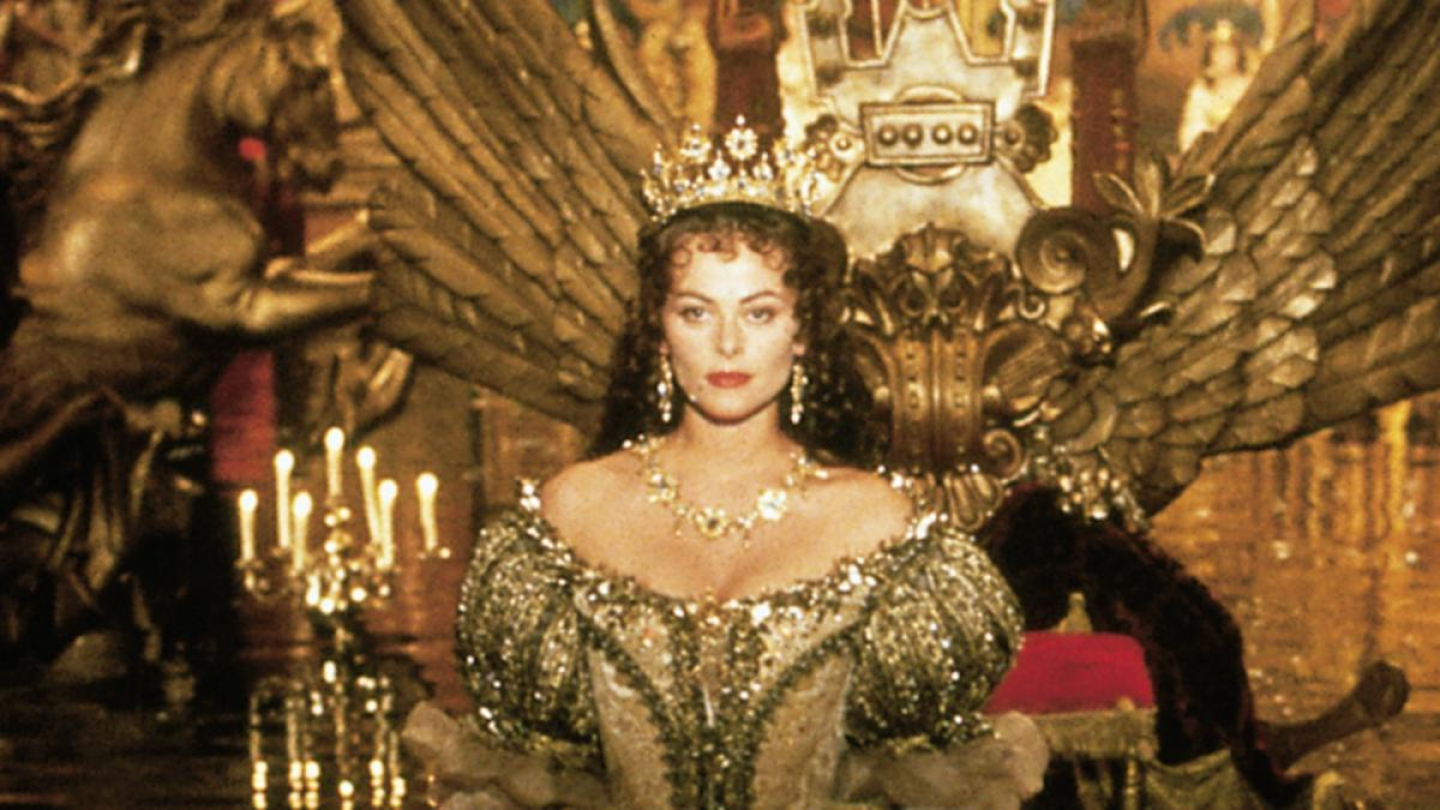 Uma mulher vestida como rainha, com coroa e trajes luxuosos, está sentada em um trono dourado ornamental em um ambiente palaciano