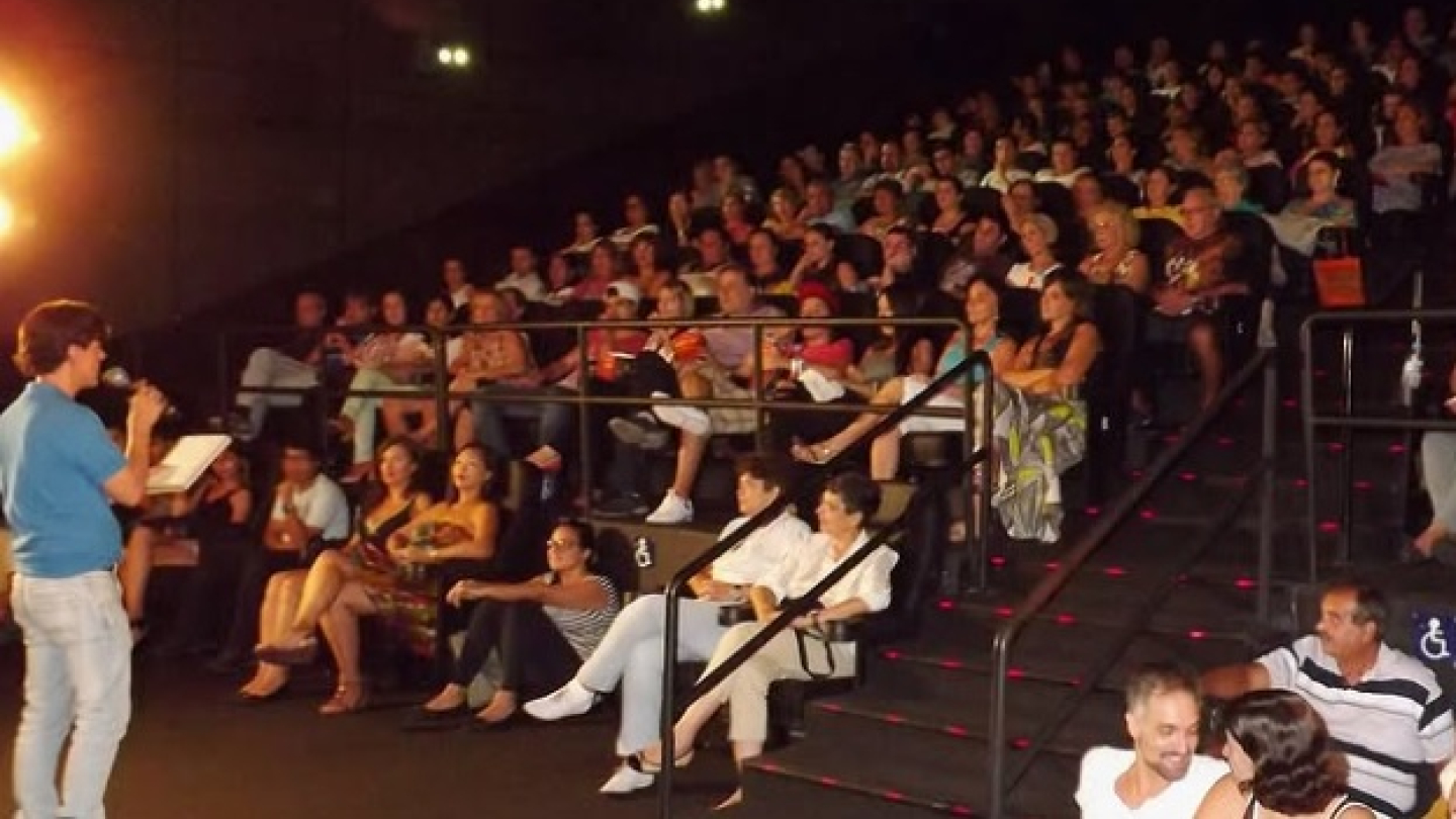 A imagem registra uma palestra de Waldemar Lopes sobre o Oscar ocorrendo em uma sala de cinema lotada, o Cine Roxy 5, no dia 10 de março