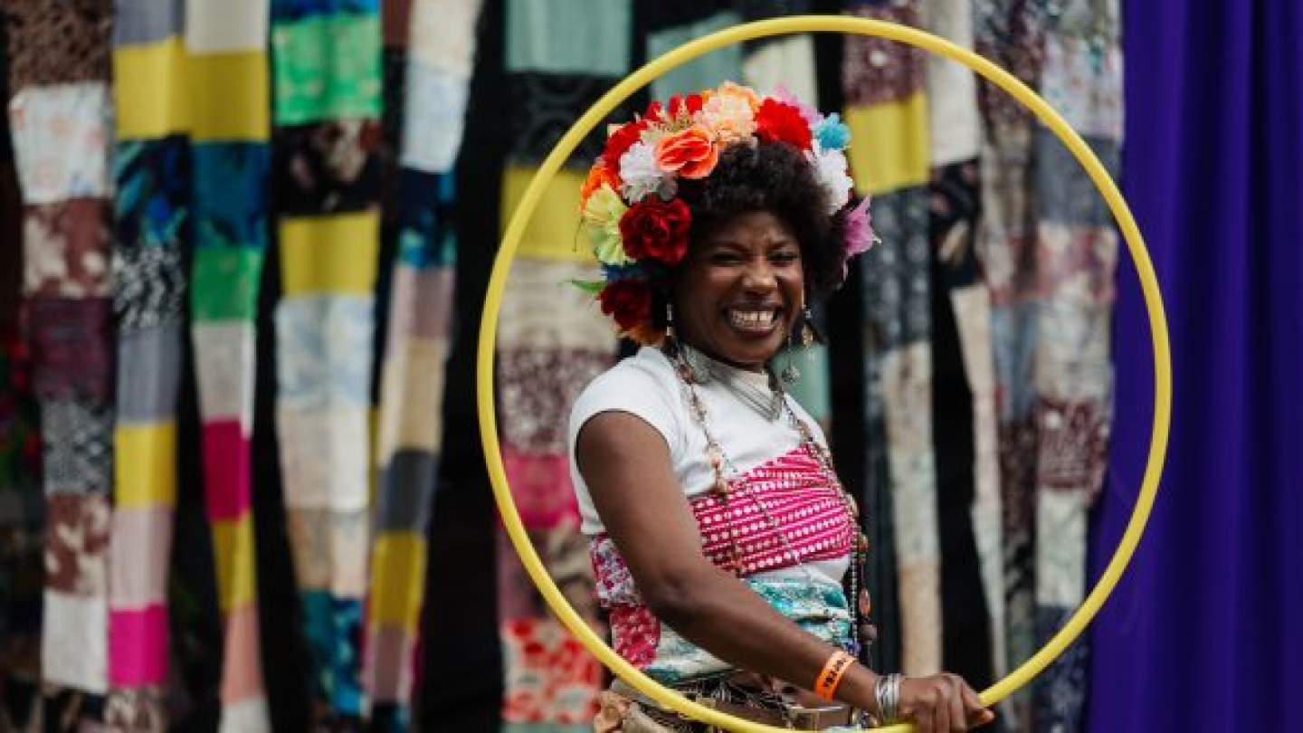 Uma artista sorridente com um adereço floral na cabeça segura um bambolê amarelo em frente a um cenário colorido feito de retalhos de tecido