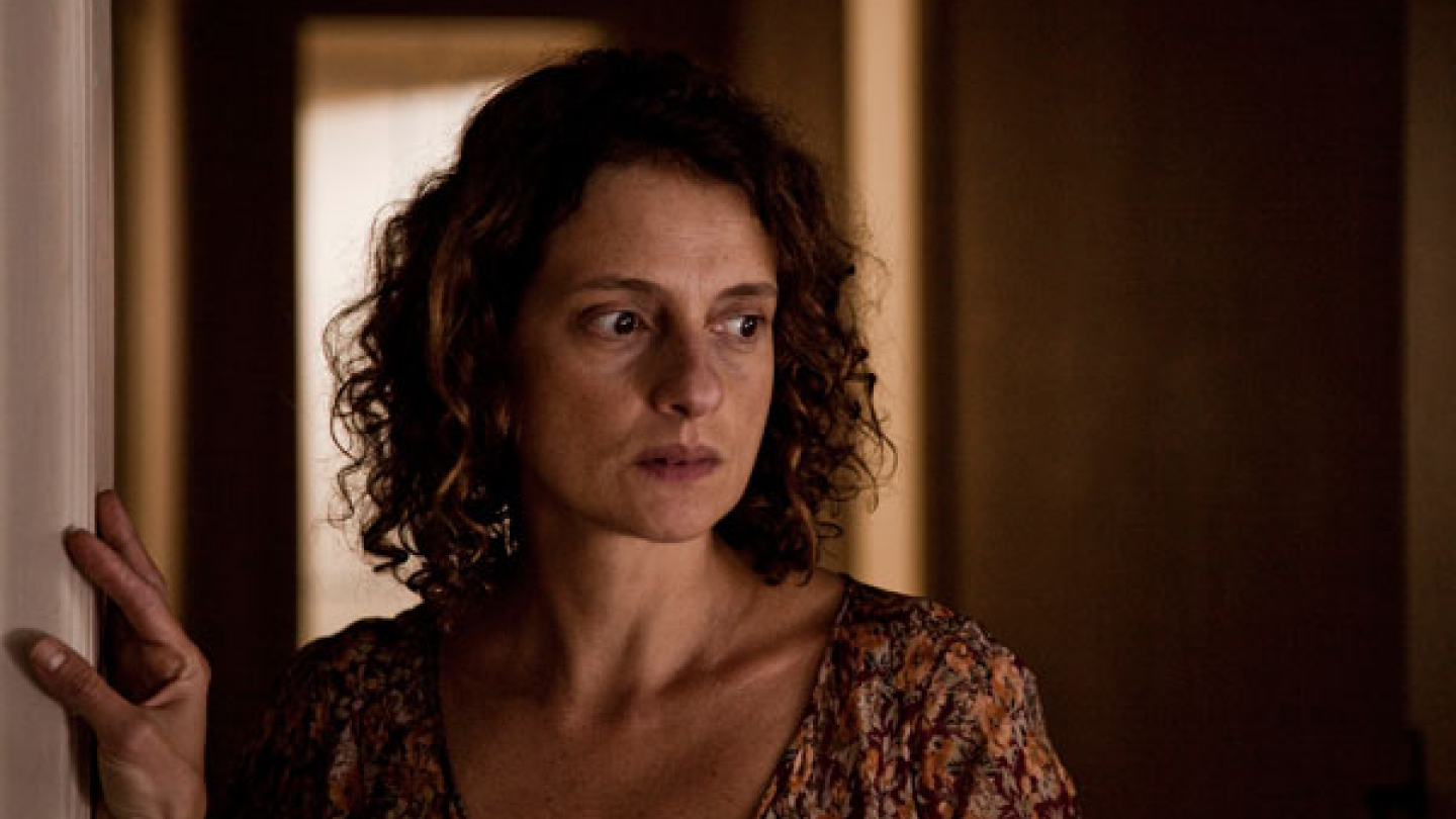 A atriz Denise Fraga aparece em uma cena dramática com expressão apreensiva e olhar perdido em um ambiente doméstico pouco iluminado