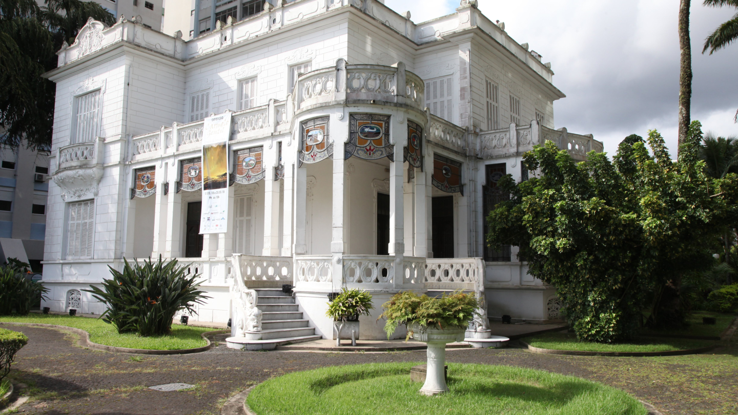 Fachada de um casarão histórico branco com detalhes arquitetônicos clássicos e vitrais, cercado por um jardim com gramado e arbustos