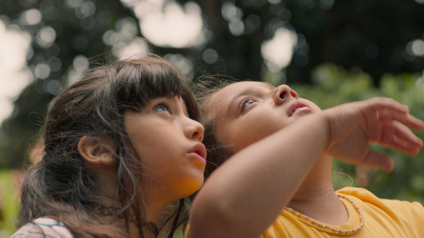 Duas meninas olham com curiosidade e admiração para cima, em meio a um cenário natural com iluminação suave