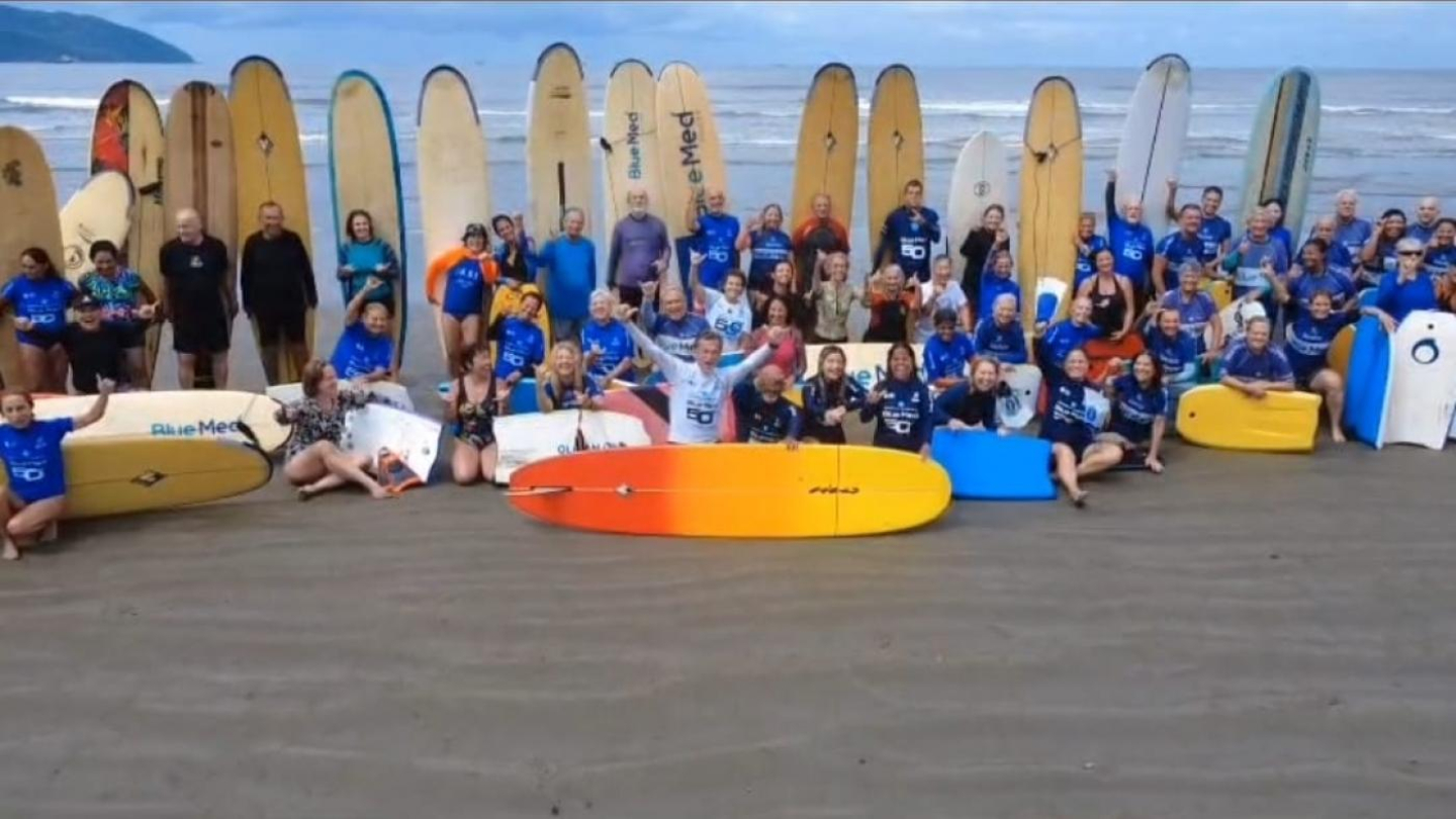 Uma grande e animada comunidade de surfistas de diversas idades posa reunida em uma praia, segurando seus pranchões e bodyboards à beira-mar