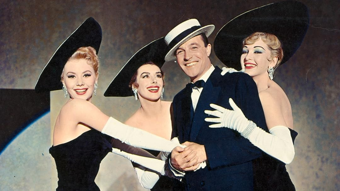 Cena clássica do filme musical "Les Girls", mostrando o ator Gene Kelly sorridente e elegante ao lado de três dançarinas em figurinos pretos sofisticados com grandes chapéus