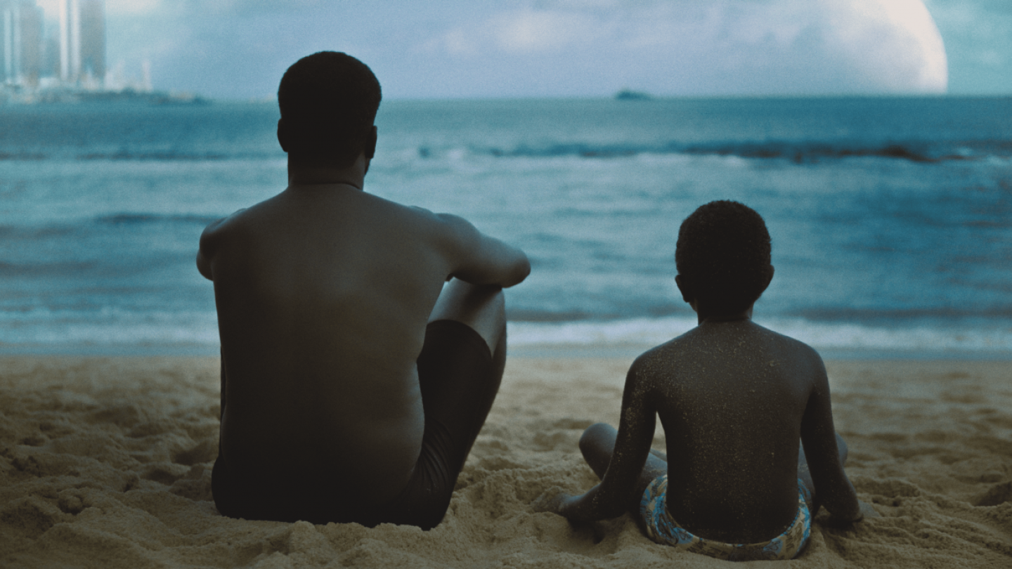 Um homem e uma criança negra estão sentados de costas na areia, observando o mar sob um céu nublado com prédios ao fundo