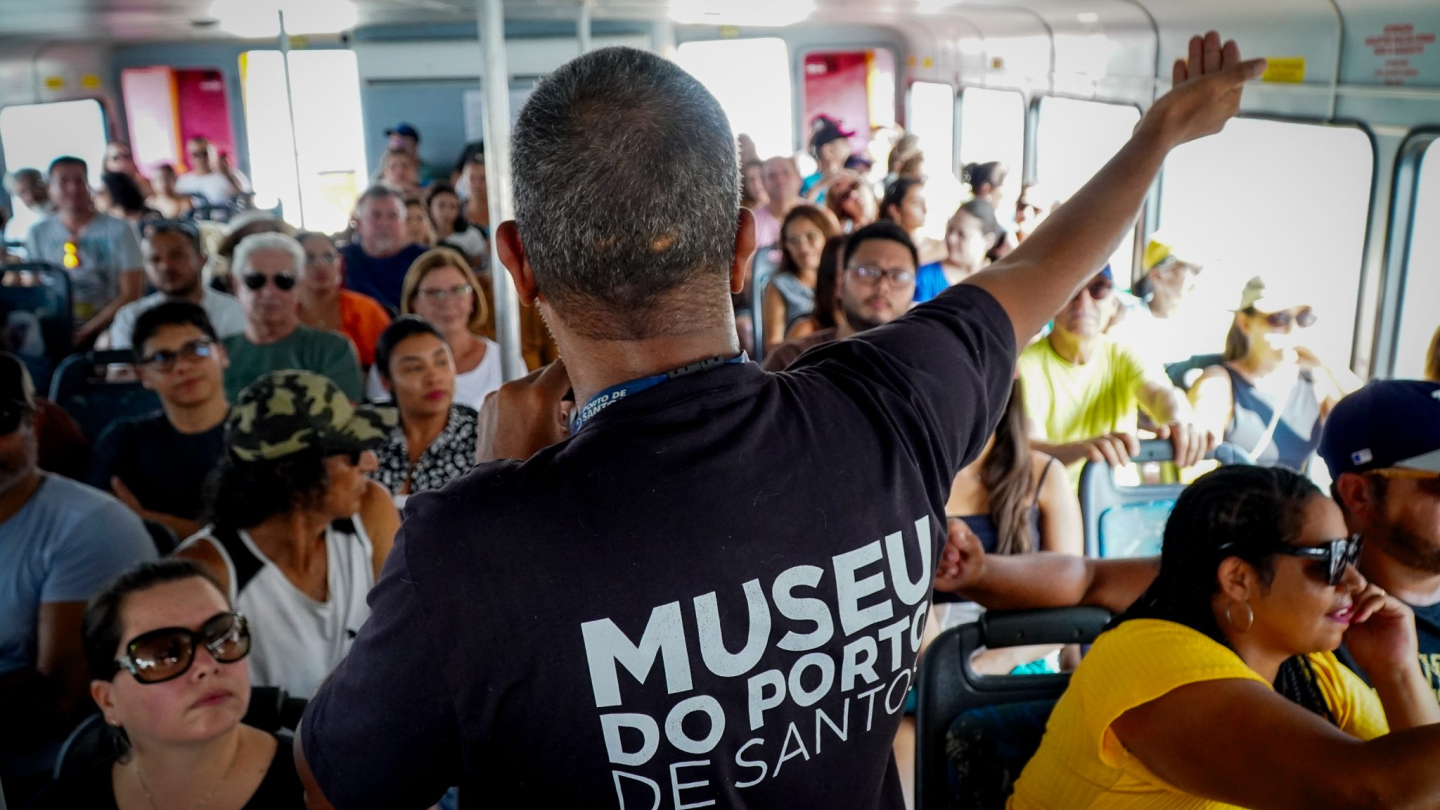 Um guia de costas, vestindo uma camiseta do Museu do Porto de Santos, gesticula para um grupo de passageiros atentos durante um passeio de barco