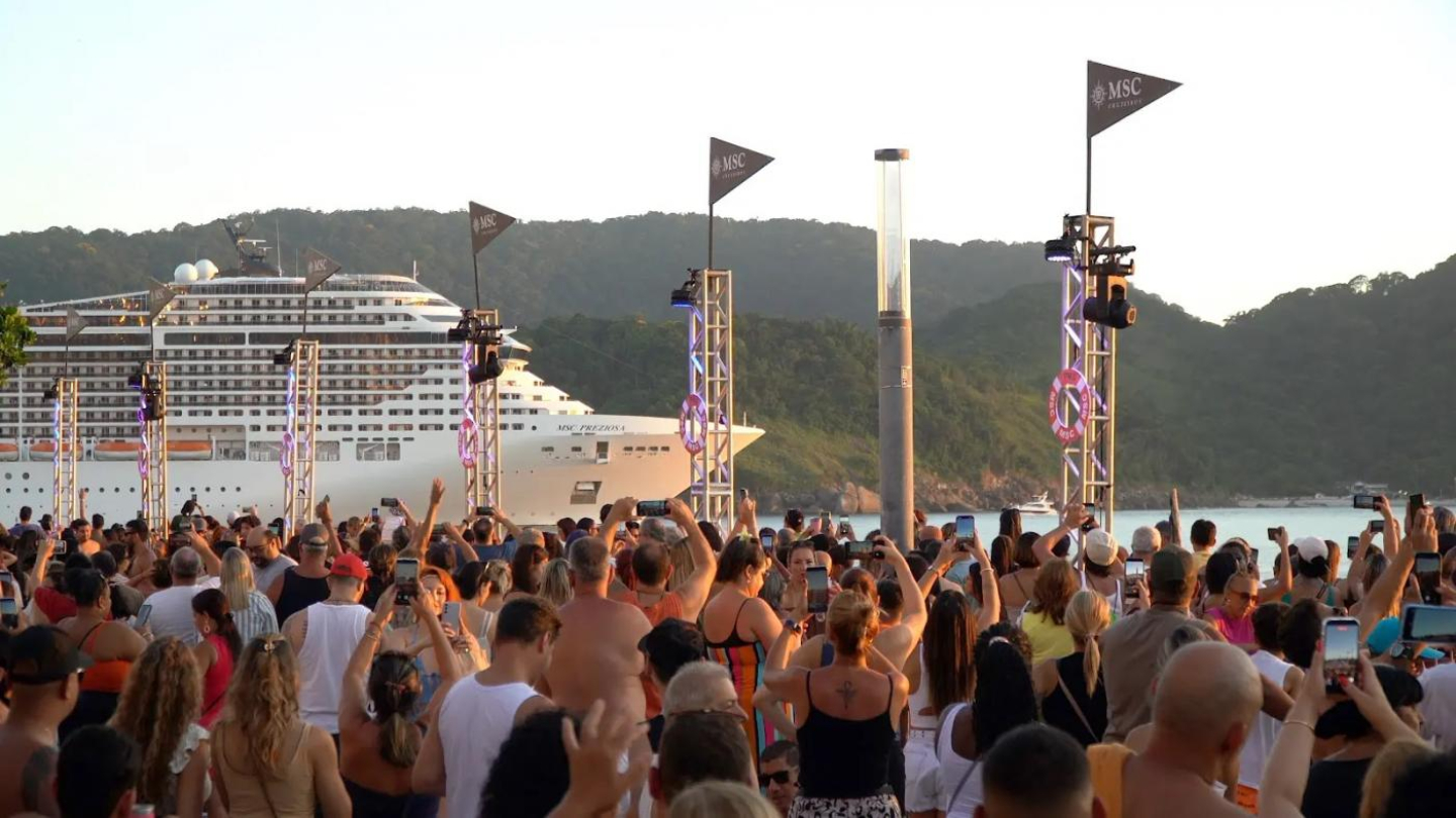 Evento repleto de pessoas com navio de cruzeiro passando ao fundo