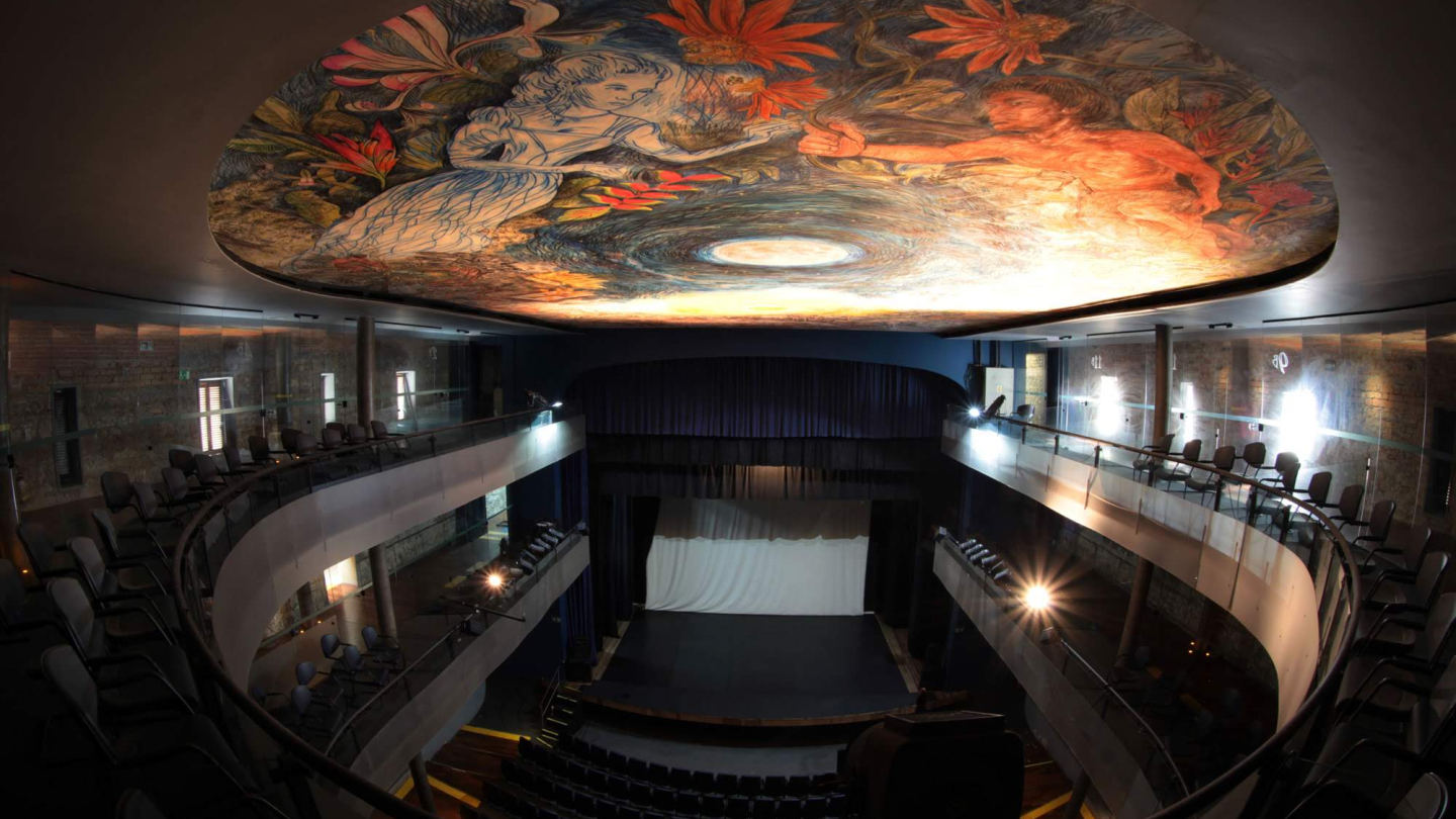 Visão interna do Teatro Guarany, destacando a plateia em ferradura e o teto adornado por uma pintura artística vibrante que retrata figuras humanas e elementos da natureza