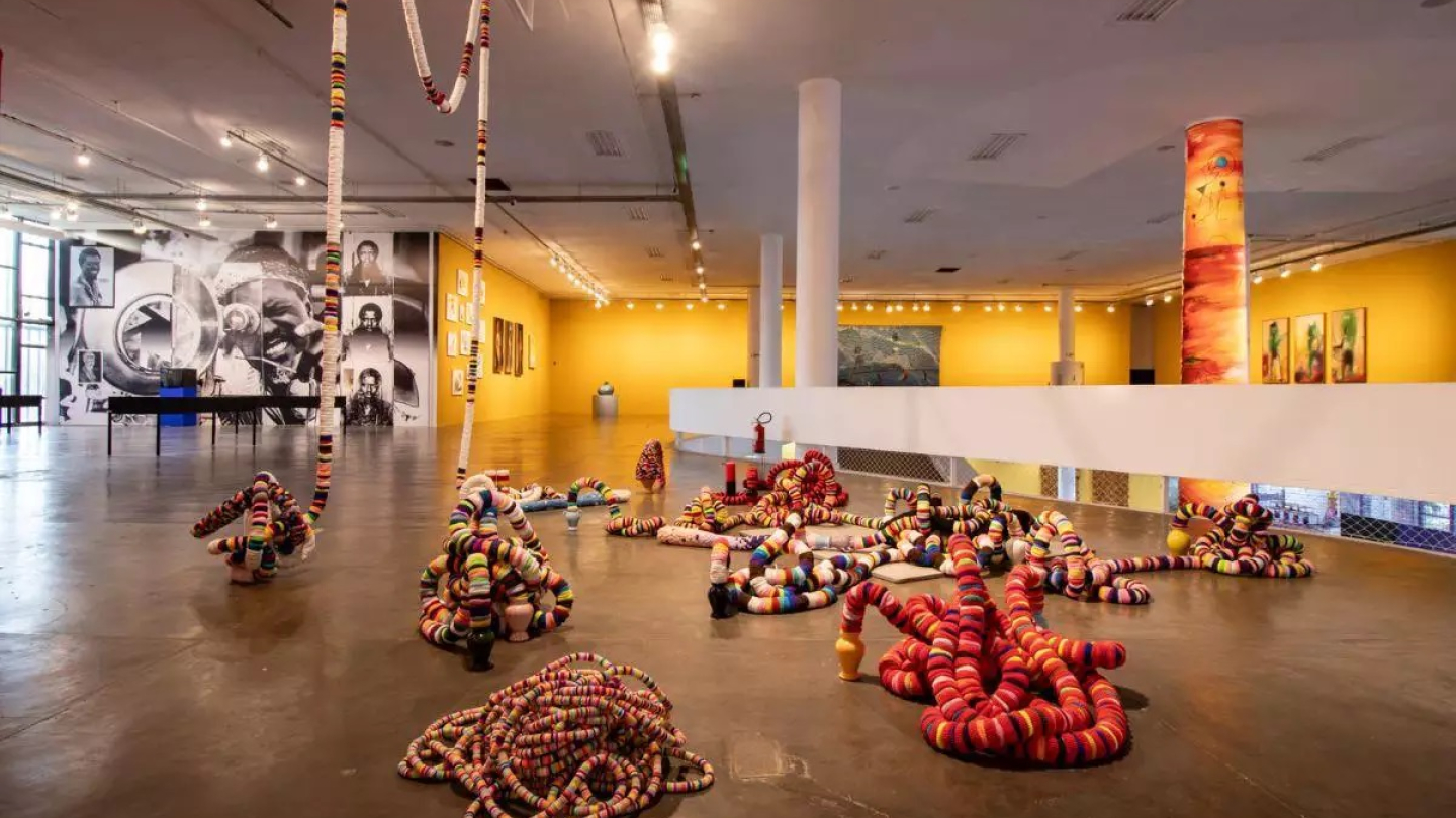 Exposição de arte contemporânea no Sesc apresentando esculturas maleáveis e coloridas que se espalham pelo chão e pendem do teto, em um amplo salão
