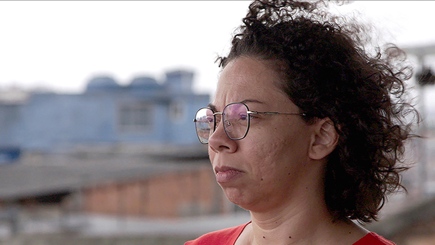 Retrato em close de uma mulher de óculos e cabelos cacheados, com expressão serena e fundo urbano levemente desfocado