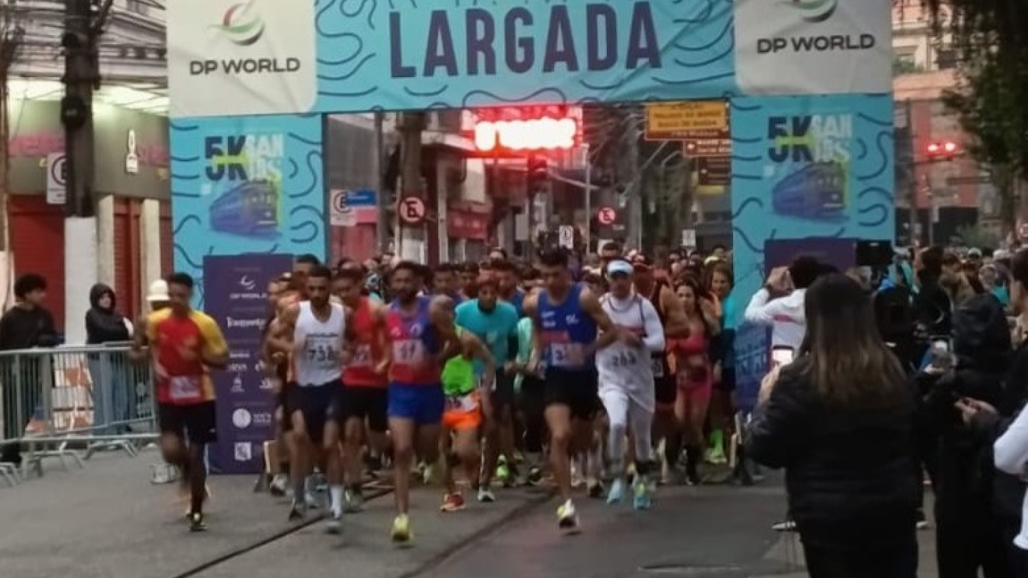 Corredores cruzam a linha de largada de uma prova de rua em Santos, sob um pórtico azul, em uma via com trilhos de bonde