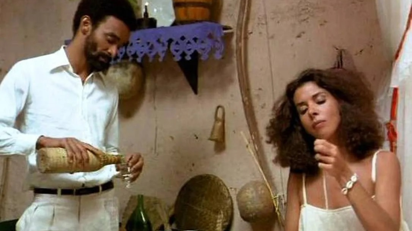 Cena do filme O Cortiço mostrando um homem servindo uma bebida enquanto uma mulher, em primeiro plano, observa com expressão pensativa em um ambiente rústico