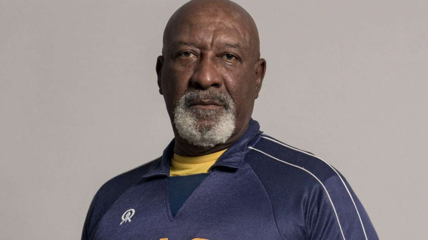 Retrato de um homem negro de barba grisalha vestindo uma camisa esportiva azul da seleção brasileira de vôlei e segurando uma bola
