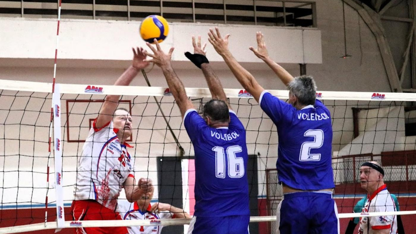 Jogadores de vôlei da categoria master disputam um lance junto à rede, com um atacante de uniforme branco e vermelho saltando contra o bloqueio de dois jogadores de uniforme azul