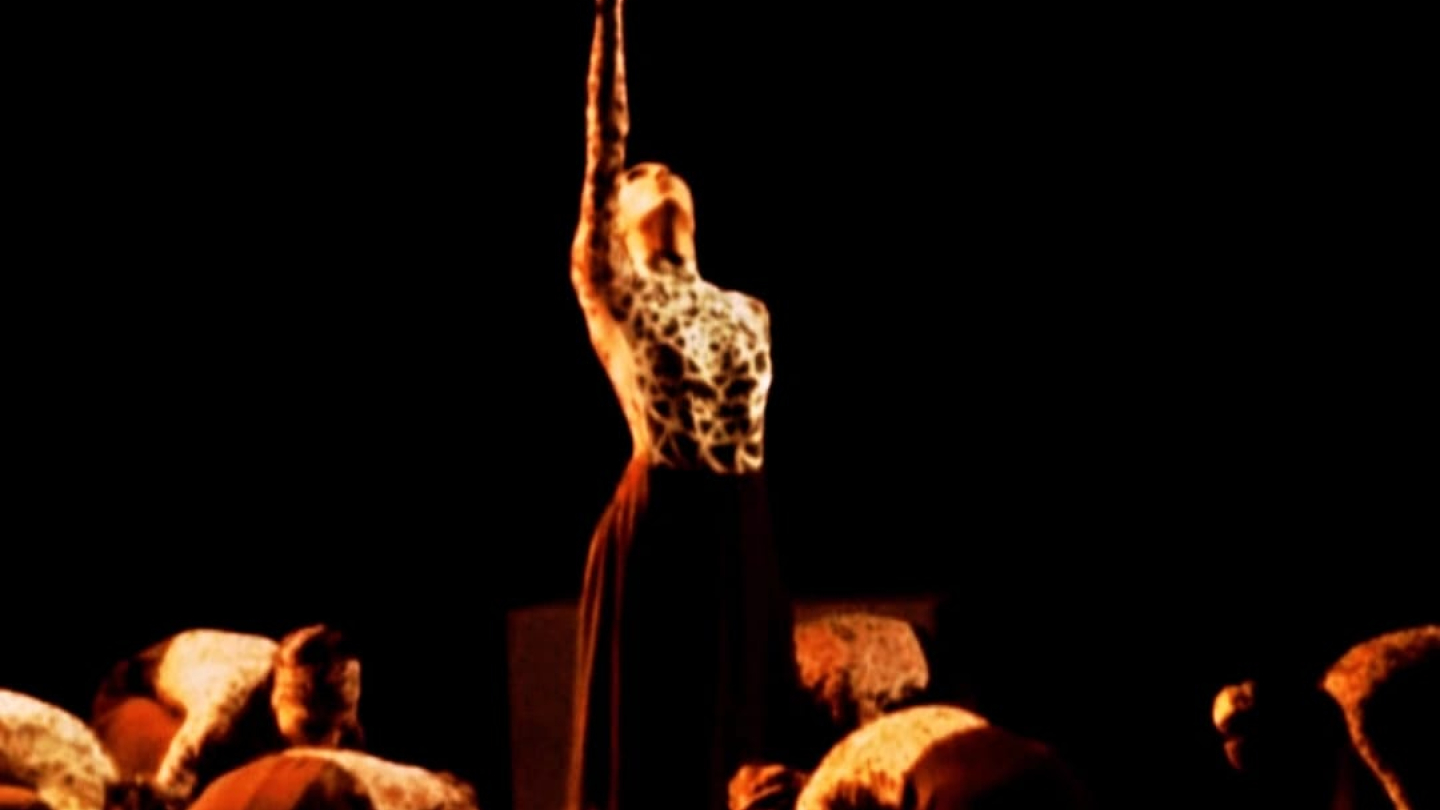 Uma bailarina em pose vertical com braço elevado destaca-se no centro de um palco escuro, cercada por outras dançarinas agachadas e curvadas ao chão