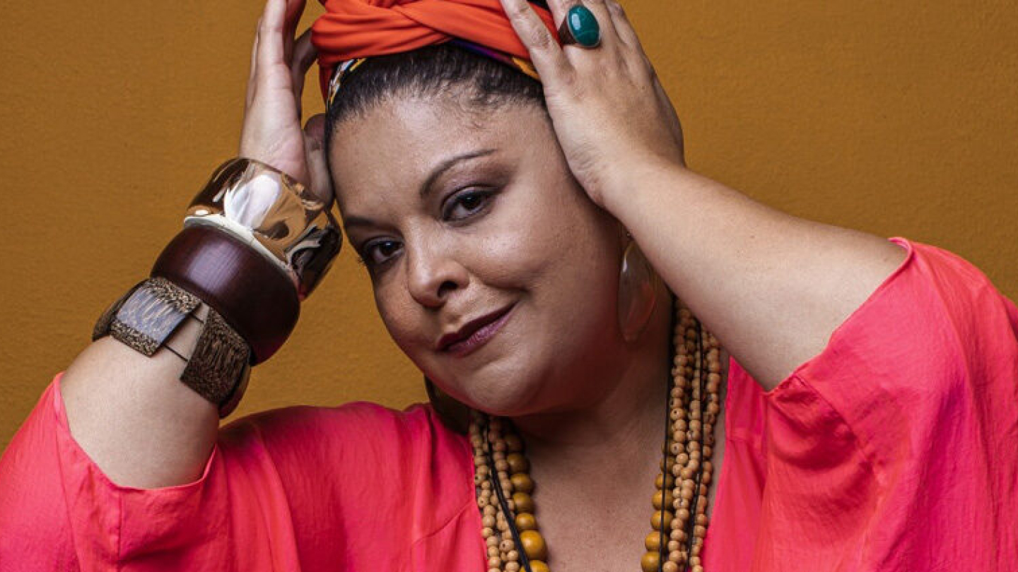 Uma mulher negra posa inclinando a cabeça levemente, vestida com roupas e turbante em tons de rosa e laranja, adornada com pulseiras e colares grandes contra um fundo amarelo