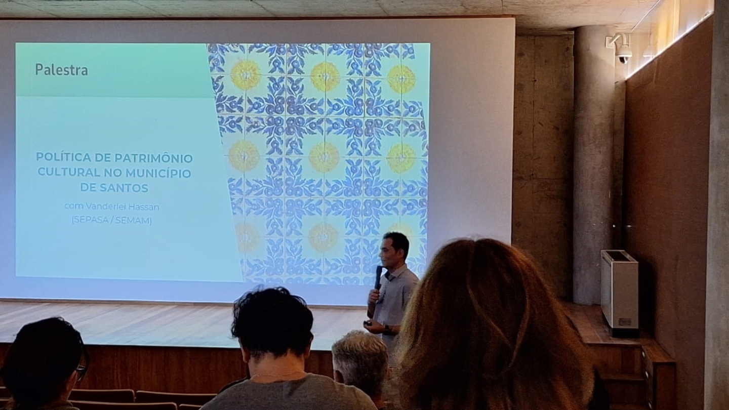 Um palestrante de camisa cinza fala ao microfone em um palco diante de uma plateia, projetando um slide sobre a "Política de Patrimônio Cultural no Município de Santos"