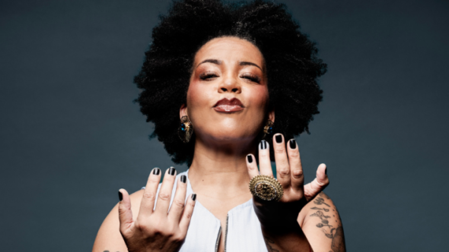 Uma mulher negra com cabelo black power e unhas pintadas de preto posa com as mãos abertas à frente do corpo contra um fundo cinza escuro