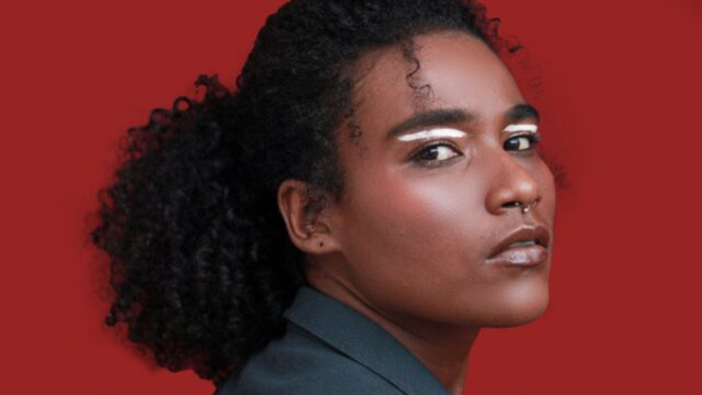 Uma mulher negra, vista de perfil com o rosto virado para a câmera, exibe cabelos cacheados presos e uma maquiagem marcante com traços brancos nas sobrancelhas contra um fundo vermelho